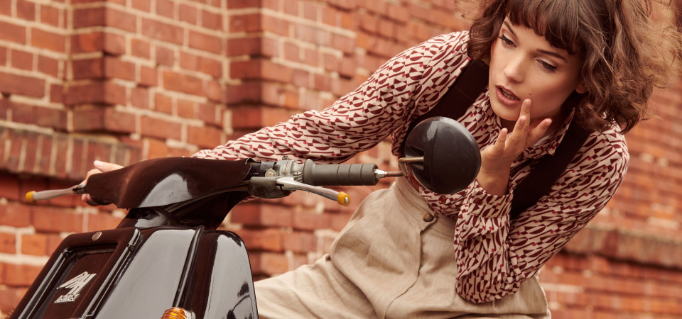 Vintage-Hosen Eine Frau in einer Vintage-Bluse und einer hellen Marlenehose sitzt auf einem Motorroller