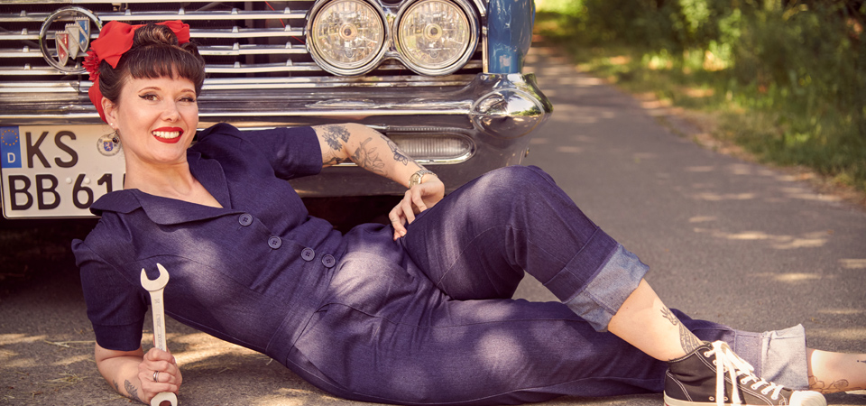 Jumpsuits und Overalls Eine Frau posiert im Jeans-Overall vor einem Oldtimer-Auto und hält einen Schraubenschlüssel in der Hand