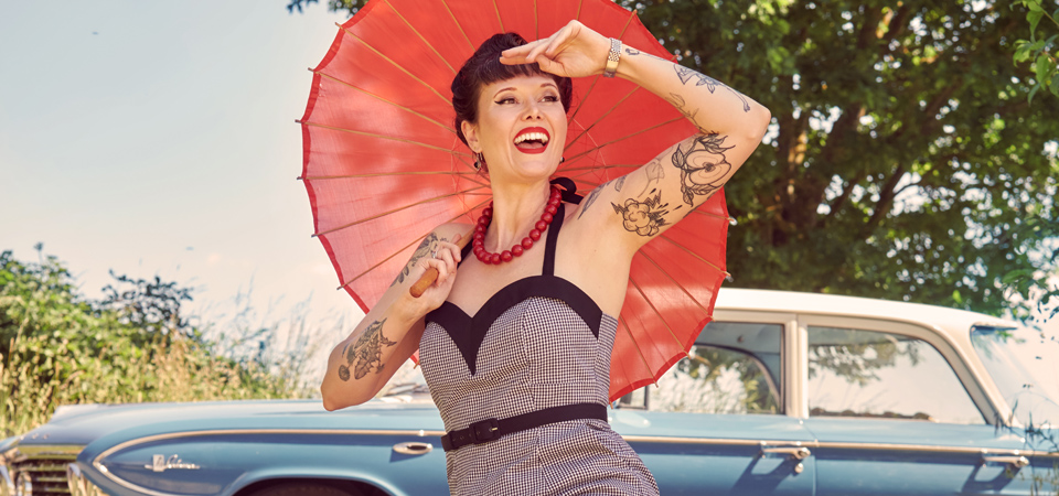 Playsuits im Vintage-Stil Eine Frau in einem karierten Playsuit hält einen Sonnenschirm und sitzt auf der Wiese vor einem Oldtimer-Auto